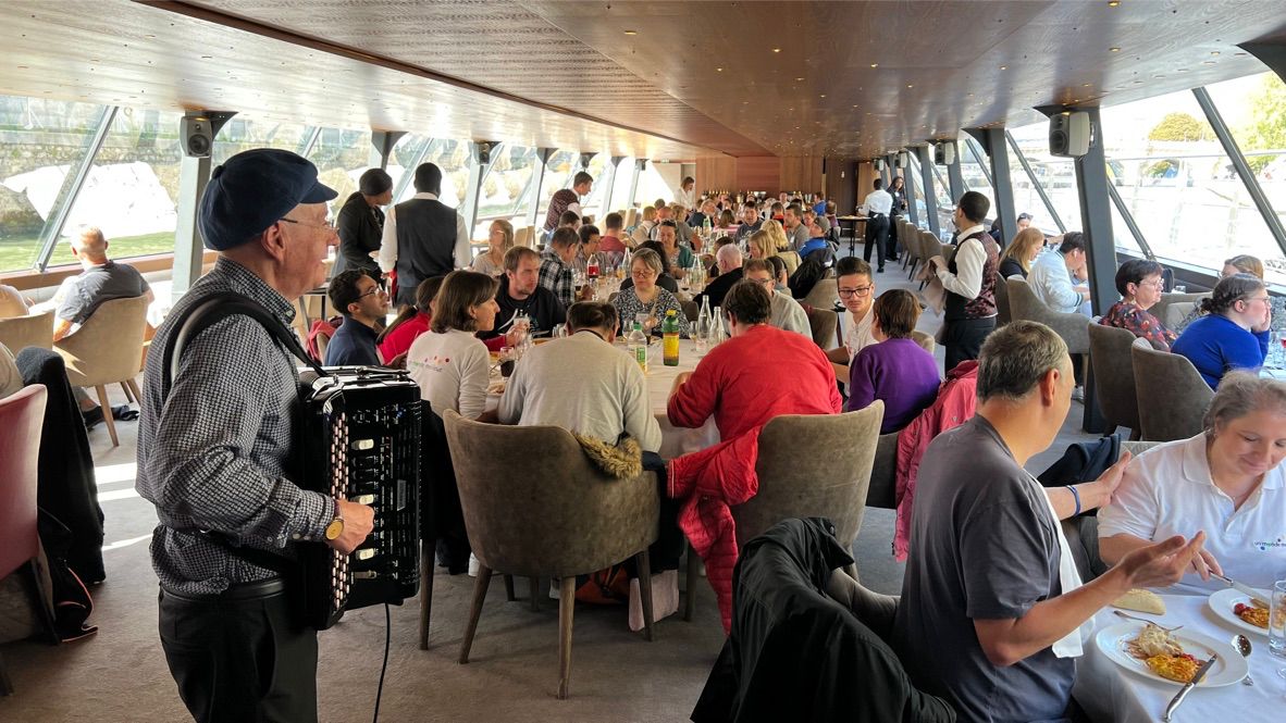 Un accordéoniste joue pendant qu’un grand groupe partage un repas sur un bateau.